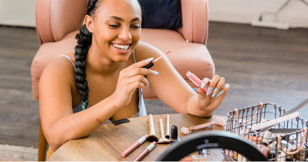 Smiling woman sitting at a table applying lip gloss while recording a beauty video, with makeup brushes and products spread out in front of her