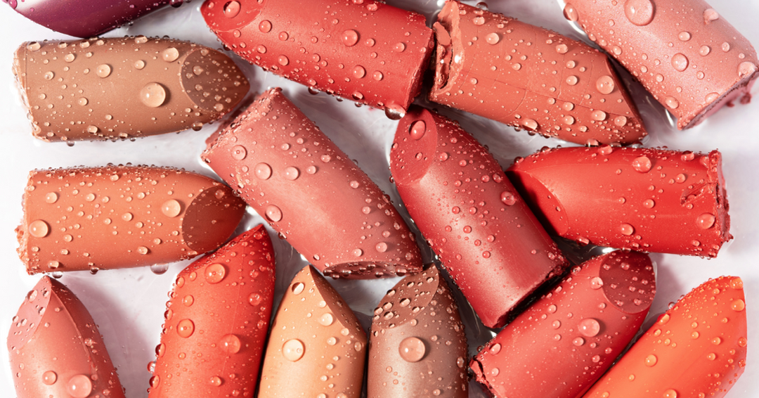 A close-up image of various lipstick bullets in shades of nude, pink, coral, and red, arranged haphazardly on a white surface. Each lipstick is covered in water droplets, giving them a fresh and dewy appearance