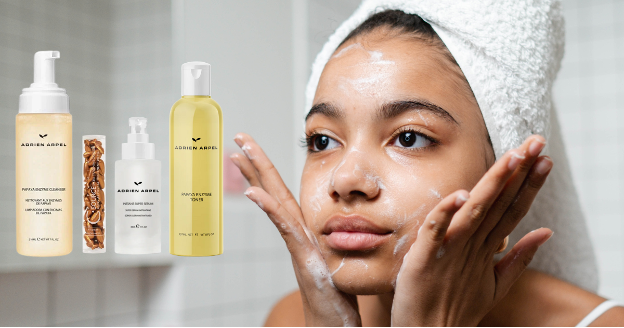 A woman with a towel wrapped around her head washes her face with a foaming cleanser. Beside her are product images of Adrien Arpel's Papaya Enzyme Cleanser, Papaya Enzyme Toner, C Source Vitamin C capsules, and Instant Super Serum.